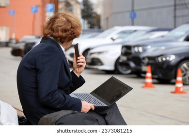 Man sitting with a laptop and smartphone on a bench on city street on parked cars background - Powered by Shutterstock