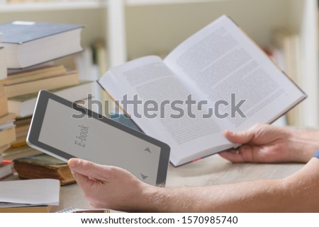 Man sitting in a library and holding in hands a modern ebook reader and paper books, bookshelf in the background 