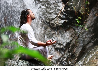 Man Sitting In Meditation Yoga On Rock At Waterfall In Tropical
