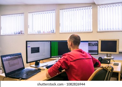 A Man Is Sitting At The Office Behind A Worker's Desk. The Engineer Monitors The Computer Network. A Specialist Works For Several Computers.