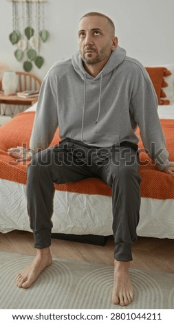 Man sitting on bed touching neck and wincing, wearing hoodie, barefoot on rug beside orange bed throw in a building bedroom; discomfort fatigue.