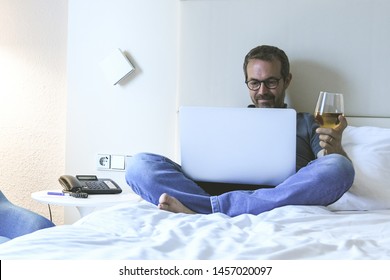 Man Sitting On A Bed Video Communicate With Remote Friends During A Trip. Smiling Male Alone In An Hotel Room Chat With Family Using Laptop And Drinking Wine. Communication Relax And Happiness Concept