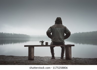 A man is sitting on a bench and looking at the lake. Back view. Also a coffee maker and a mug on the bench. Foggy morning