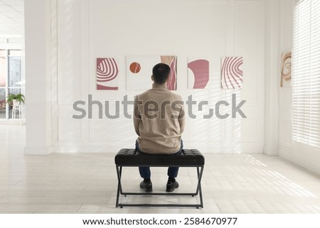 Man sitting on bench and looking at paintings in art gallery, back view