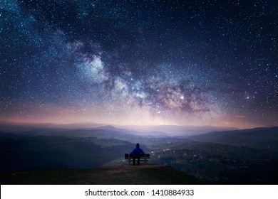 A man sitting on a bench staring at a starry sky with a Milky Way and a mountain landscape - Shutterstock ID 1410884933