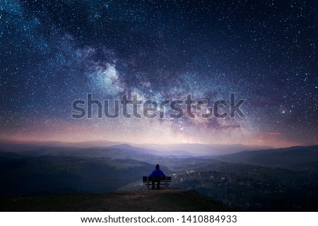 A man sitting on a bench staring at a starry sky with a Milky Way and a mountain landscape