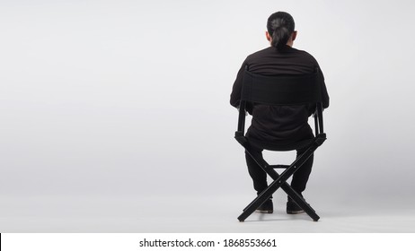 A Man Is Sitting On Black Director Chair.It Is White Background.
