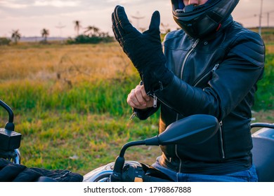 Man sitting on a black motorcycle, dressed in jeans and a black jacket, putting on black gloves with the landscape in the background.