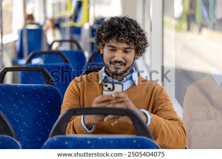 Man sitting on bus using smartphone with a smile. Casual setting in public transport with blue seats and window light. Engaged and relaxed moment captured.