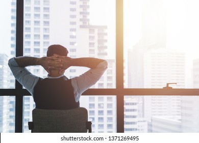 Man Sitting On The Chair With Hands Behind His Head Looking Outside Through The Window Indoors. Successful Business Entrepreneur, Achievement, Relaxation Concept.  