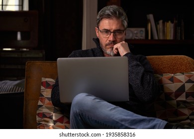 Man sitting on couch at home with laptop computer. Businessman working in home office. Portrait of mature age, middle age, mid adult man, bearded, glasses, authentic look.