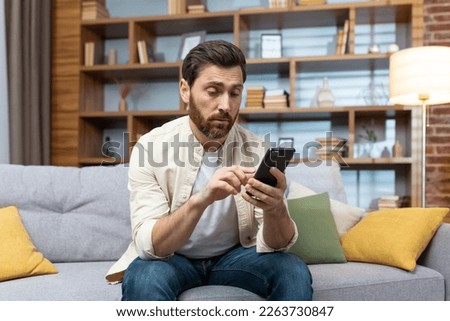 A man sitting on a couch at home, looking bored as he scrolls through dating apps on his phone. The casual setting and relaxed posture create a relatable image of modern dating culture