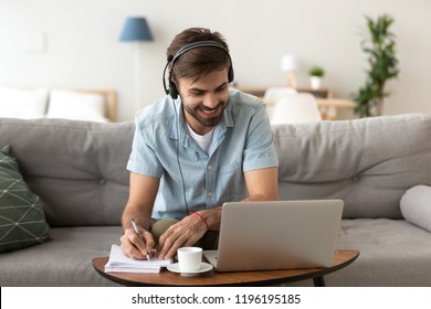 Man sitting on couch in living room at home enjoying studying using laptop and headset looking at device screen listening audio making some notes. Male has lesson online e-learning in internet concept
