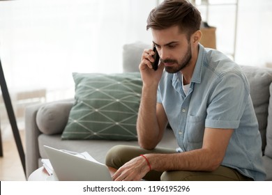 Man Sitting On Couch Reading Message Email Online Looking At Laptop Talking By Mobile Phone. Male Working At Home Has Serious Conversation With Client. Businessman Solve Problems And Matters By Phone