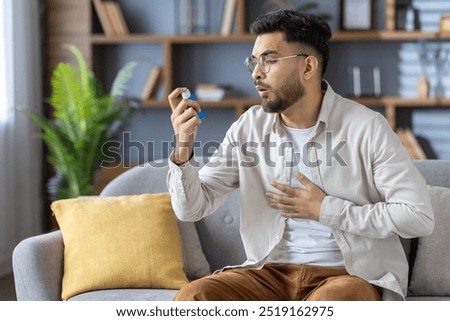 Man sitting on couch using inhaler for respiratory relief, holding chest due to breathing issue. Wearing casual clothing, looking at inhaler to alleviate discomfort