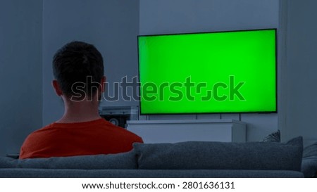 Man sitting on a couch watching a bright green screen tv in a dark living room from behind