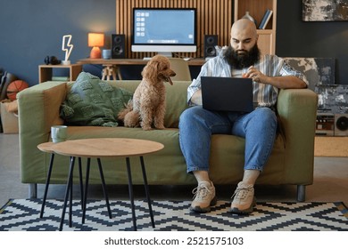Man sitting on couch working on laptop while dog stands next to him offering company in cozy living room full of modern technology and decor including speakers, books, computer monitor