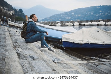 Man sitting on a dock by a marina, looking contemplative. Mountains in the background with moored boats, calm water, and a peaceful atmosphere. - Powered by Shutterstock