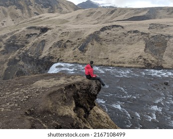 Man Sitting On Edge Of Cliff By Waterfall In Iceland 