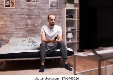 Man Sitting On The Edge Of His Bed At Night Watching Tv Holding Remote Control.