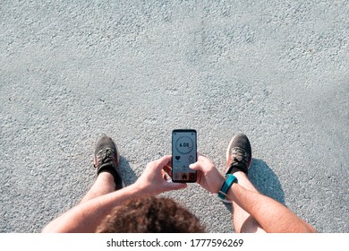 Man Sitting On The Ground Looking At An App On His Cell Phone After Running Outside. Focus On The Mobile Phone.