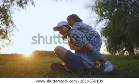 A man is sitting on the ground while a girl joyfully rides on his back. a sunset behind them, casting a warm glow across the grassy field.