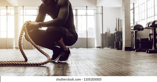 Man Sitting On His Toes Holding A Pair Of Battle Ropes For Workout. Guy At The Gym Working Out With Fitness Rope.