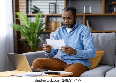 Man sitting on living room sofa reading letter with concerned expression. Home office setup with laptop and paperwork on table. Concept of financial stress and concern. - Powered by Shutterstock