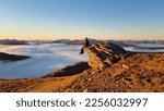 Man sitting on a mountain top looking over a cloud inversion in th valleys with mountain tops poking out while the sun sets