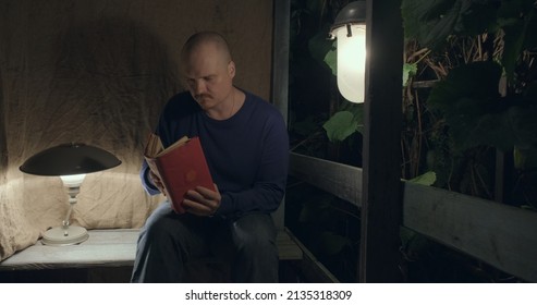 Man Is Sitting On An Old Village Porch. He Picks Up An Old Book. Dark Night, Table Lamp And A Retro Lantern Are Lit. The Concept Of Hobbies, Antique Books And Vintage Items