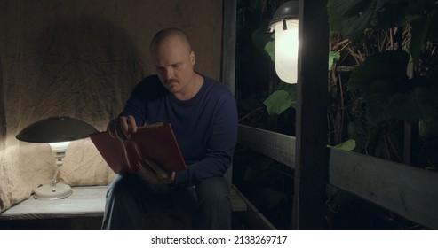 Man Is Sitting On An Old Village Porch. He Picks Up An Old Book. Dark Night, Table Lamp And A Retro Lantern Are Lit. The Concept Of Hobbies, Antique Books And Vintage Items