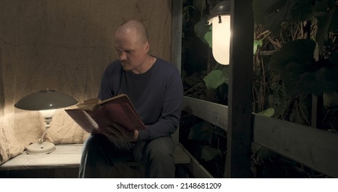 Man Is Sitting On An Old Village Porch. He Picks Up An Old Book. Dark Night, Table Lamp And A Retro Lantern Are Lit. The Concept Of Hobbies, Antique Books And Vintage Items