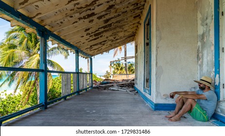 Man Sitting On The Patio Of An Old Decayed House On A Tropical Beach.
