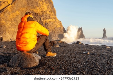 A man sitting on the Reynisfjara Black Sand Beach looking at the beach with a wave - Powered by Shutterstock