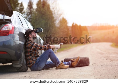 The man is sitting on the road by the car in the nature
