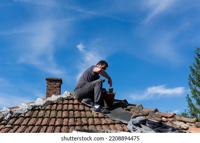 The man sitting on the roof of an old house and repairing tile with cement in the village on a sunny day. Blue sky in the background