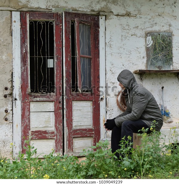 Man sitting on sink at\
the porch of an abandoned house with old door frame window and\
smudged mirror.