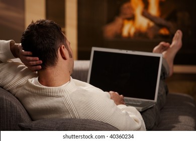 Man Sitting On Sofa At Home In Front Of Fireplace And Using Laptop Computer, Rear View.