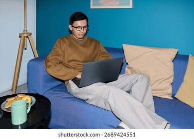 Man sitting on sofa using laptop, wearing glasses and focusing on screen, working or studying in modern home setting with snacks and drink on table