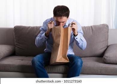 A Man Sitting On Sofa Vomiting Into Paper Bag At Home