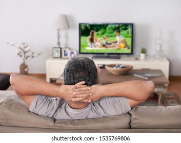 Man Sitting On A Sofa Watching Tv With Hands Folded Behind His Head