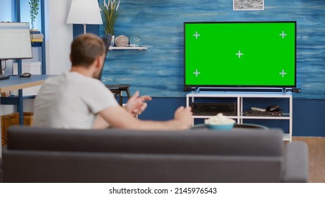 Man Sitting On Sofa Watching Sport Game On Green Screen Tv Mockup Encouraging Favourite Team While Having A Good Time. Sports Fan Relaxing Looking At Television With Chroma Key Display In Living Room.