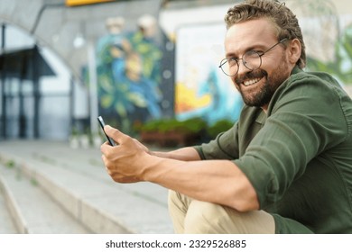 Man sitting on stone stairs on street, using pocket PC. With compact and portable device in hand, he effortlessly combines technology with surroundings. urban backdrop adds modern touch to scene. High - Shutterstock ID 2329526885