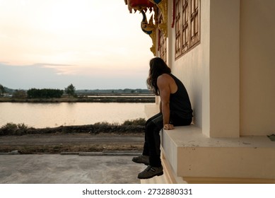 Man sitting on a temple ledge overlooking a calm river during sunset