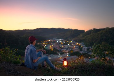 Man Sitting On Top Of A Mountain With A Kerosene Lamp Burning By His Side And Lights Of A City At Night In The Background. Energy And Electricity Concept.