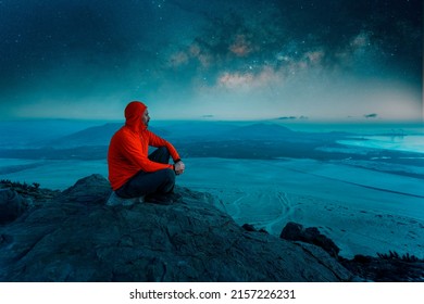 Man Sitting On Top Of A Mountain Contemplating The Horizon Over The Ocean At Night