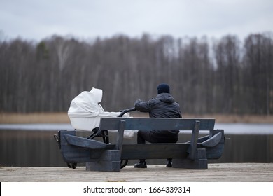 Man sitting on wooden bench and staring at lake in cold day. White baby stroller beside young father. Thinking about life. Back view. Peaceful atmosphere in nature.  - Powered by Shutterstock