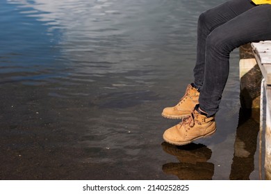 Man Sitting On Wooden Dock Above Sea. 
His Feet Dangle From Jetty. He Wears Old Brown Boots. Only Legs Is Seen In Photo. Unrecognizable Person.  