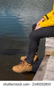 Man Sitting On Wooden Dock Above Sea. 
His Feet Dangle From Jetty. He Wears Old Brown Boots. Only Legs Is Seen In Photo. Unrecognizable Person.  Vertical Photo.