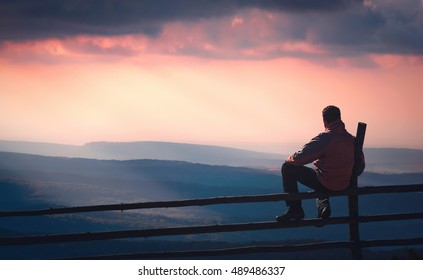 Man sitting on a wooden fence and enjoy rays of sunlight, shining through the clouds to the hills. Carpathian mountain valley.